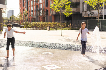 Wall Mural - Cheerful young teen girl in city fountain, girl in wet clothes is having fun and enjoying the cool summer water, background city architecture.