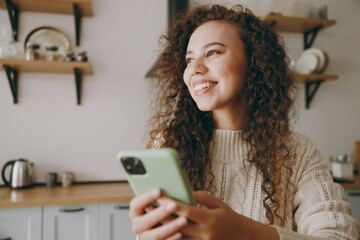 Young minded happy housewife woman wearing casual clothes sweater hold use mobile cell phone drink coffee look for recipe sit at table in light kitchen at home alone. Lifestyle cooking food concept.