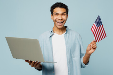 Wall Mural - Young IT man of African American ethnicity he wear shirt casual clothes hold American flag use work on laptop pc computer hold use work on laptop pc computer isolated on plain pastel blue background.