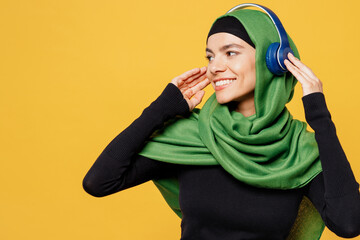 Wall Mural - Young arabian asian muslim woman wearing green hijab abaya black clothes listen to music in headphones look aside isolated on plain yellow background People uae middle eastern islam religious concept