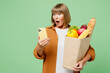 Elderly amazed woman wearing brown shirt casual clothes holding shopping paper bag with food products hold in hand use mobile cell phone isolated on plain green background. Delivery service from shop.