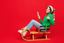 Full Body Young Fun Woman Wear Green Turtleneck Santa Hat Posing Sit Ride Sled Hold Use Mobile Cell Phone Show Thumb Up Isolated On Plain Red Background. Happy New Year 2024 Christmas Holiday Concept.