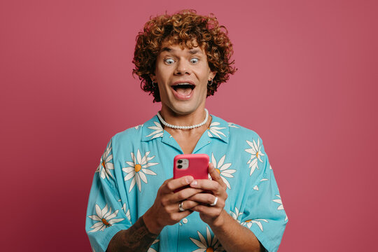 Surprised young curly hair man in funky shirt looking at his smart phone against pink background