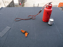 Red Propane Gas Cylinder With Connected Blowtorch Standing On Almost Finished New Bitumen Roofing Felt Covered Flat Rooftop.