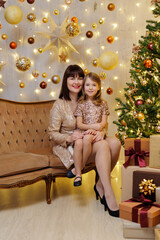 Wall Mural - Mother and little daughter in golden dresses sitting on sofa in christmas decorations