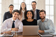 © fizkes - Happy young multiethnic team posing at workplace table, looking at camera, smiling, meeting at laptop for teamwork, successful collaboration, brainstorming on project. Business group portrait