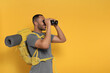 © New Africa - Happy tourist with backpack looking through binoculars on yellow background, space for text