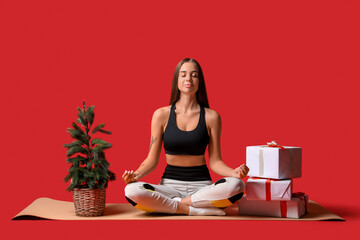 Sticker - Sporty young woman with Christmas tree and presents meditating on red background