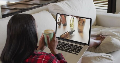 Poster - Biracial woman using laptop on couch at home online shopping for beauty products, slow motion