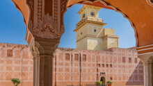 Details Of Ancient Indian Architecture. Through The Openwork Arch With A White Ornament, The Red Sandstone Wall Of The City Palace Is Visible. A Clock Tower On A Blue Sky Background. India. Jaipur.