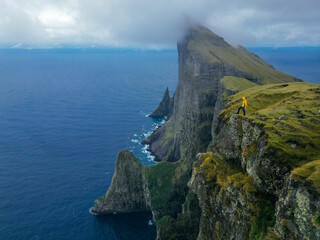 Wall Mural - A man in a yellow jacket stands at the edge of a towering cliff on the Faroe Islands, capturing the breathtaking vastness of nature and misty morning landscape. Beautiful autumn morning. Autumn hilly 