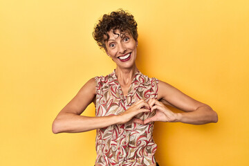 Wall Mural - Mid-aged caucasian woman on vibrant yellow smiling and showing a heart shape with hands.