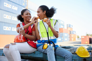 young female African factory workers or engineers having lunch and eating bread together in container warehouse storage