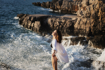 Wall Mural - Young cheerful relaxed woman in a white shirt enjoying the sea waves and splashes while standing on a stone shore