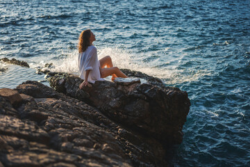 Wall Mural - Young cheerful relaxed woman in a white shirt enjoying a wonderful moment of life while sitting on the seashore at sunset