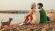 Beautiful gay couple in love talking and laughing while sitting on the beach at dawn, playing with dog