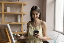 Happy Woman Using Smart Phone Distracted From Painting Standing Near Easel In Art Studio. Art-school Female Student Take Break Using Cellphone, Read Message, Check Schedule Using Mobile Application