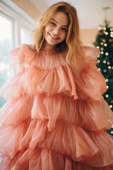 Wall Mural - A stunning woman in a flowing pink dress stands before a festive christmas tree, her radiant smile lighting up the room as she playfully runs her fingers along the ruffled fabric of her fashion