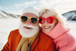 © Glittering Humanity - An eccentric senior couple embraces the winter season with funky fashion and bold eyewear, smiling under the snowy sky while sporting red goggles and sunglasses against the majestic mountain backdrop