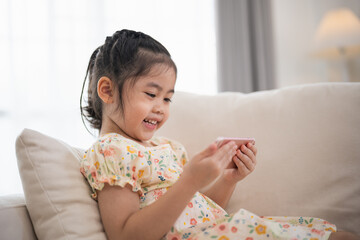 Wall Mural - Asian child girl looking using and touch mobile phone screen on couch sofa. Baby smiling funny time to use mobile phone. Too much screen time. Cute girl watching videos while tv, Internet addiction.