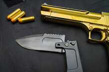 Firearms And Bladed Weapons. Gold Color Desert Eagle Pistol And Folding Tactical Knife, Close Up.