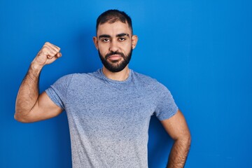 Canvas Print - Middle east man with beard standing over blue background strong person showing arm muscle, confident and proud of power