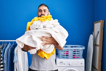 Wall Mural - Middle east man with beard holding pile of laundry puffing cheeks with funny face. mouth inflated with air, catching air.