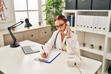 Poster - Young caucasian woman doctor talking on telephone writing on document at clinic