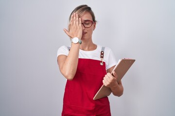 Poster - Young blonde woman wearing waiter uniform holding clipboard covering one eye with hand, confident smile on face and surprise emotion.