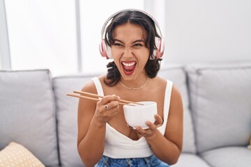 Wall Mural - Young hispanic woman eating asian food using chopsticks angry and mad screaming frustrated and furious, shouting with anger. rage and aggressive concept.