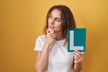 Poster - Beautiful brunette woman holding l sign for new driver serious face thinking about question with hand on chin, thoughtful about confusing idea