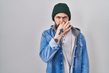 Poster - Young hispanic man with tattoos wearing wool cap smelling something stinky and disgusting, intolerable smell, holding breath with fingers on nose. bad smell