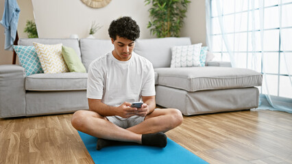 Poster - Young latin man using smartphone sitting on yoga mat at home