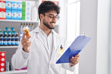 Wall Mural - Young hispanic man pharmacist holding pills bottle reading prescription at pharmacy