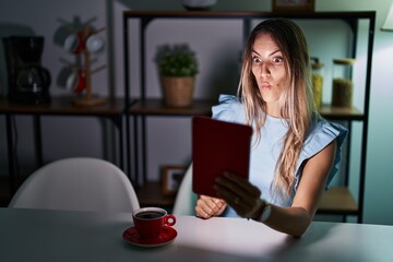 Canvas Print - Young hispanic woman using touchpad sitting on the table at night making fish face with lips, crazy and comical gesture. funny expression.