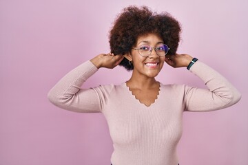 Sticker - Young african american woman standing over pink background smiling pulling ears with fingers, funny gesture. audition problem