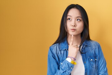 Poster - Young asian woman standing over yellow background thinking concentrated about doubt with finger on chin and looking up wondering