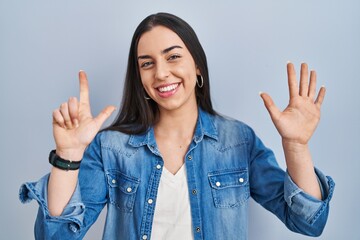 Sticker - Hispanic woman standing over blue background showing and pointing up with fingers number seven while smiling confident and happy.