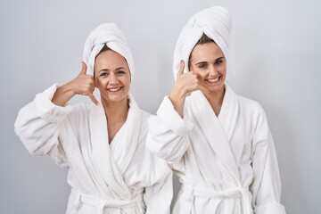 Canvas Print - Middle age woman and daughter wearing white bathrobe and towel smiling doing phone gesture with hand and fingers like talking on the telephone. communicating concepts.