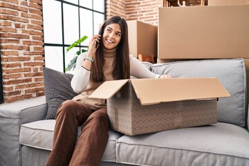 Sticker - Young beautiful hispanic woman talking on smartphone unpacking cardboard box at new home