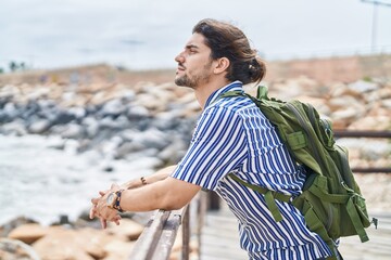 Young hispanic man tourist wearing backpack leaning on balustrade at seaside