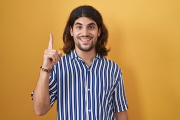 Sticker - Hispanic man with long hair standing over yellow background pointing finger up with successful idea. exited and happy. number one.