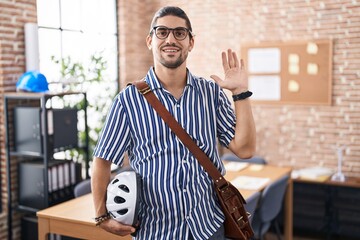 Wall Mural - Hispanic man with long hair working at the office holding bike helmet waiving saying hello happy and smiling, friendly welcome gesture