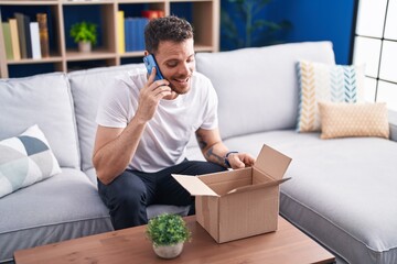 Wall Mural - Young hispanic man talking on smartphone unpacking cardboard box at home