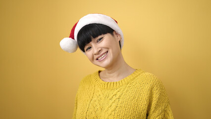 Wall Mural - Young chinese woman smiling confident wearing christmas hat over isolated yellow background