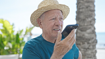 Canvas Print - Senior grey-haired man tourist wearing summer hat sending voice message by smartphone at seaside