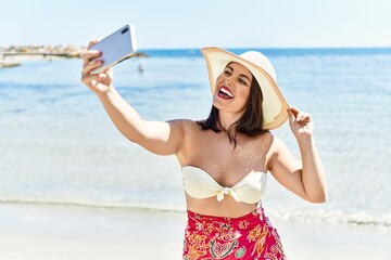 Wall Mural - Young beautiful hispanic woman tourist smiling confident make slefie by smartphone at seaside