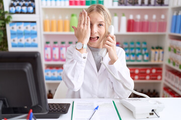 Poster - Young caucasian woman working at pharmacy drugstore speaking on the telephone covering one eye with hand, confident smile on face and surprise emotion.