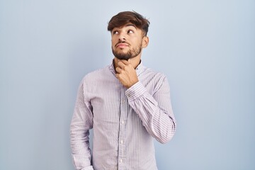 Sticker - Arab man with beard standing over blue background with hand on chin thinking about question, pensive expression. smiling with thoughtful face. doubt concept.