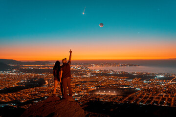 silhouettes of a couple standing on the top of the hill looking at starry night with Moon and shooting star in the sky over the city light 